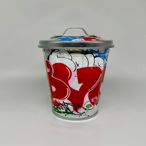 May include: A small, silver, metal trash can with a lid and a handle. The can is decorated with colorful graffiti, including the word "BYE" in large, red letters.