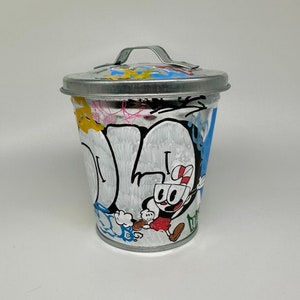 May include: A small, silver metal trash can with a handle. The can is decorated with colorful graffiti and a cartoon character. The character is wearing a red and white hat and a red shirt. The character is holding a cup in one hand and a spoon in the other.