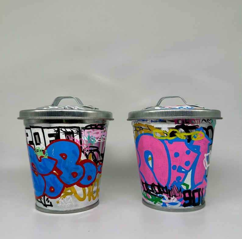 May include: Two silver trash cans with lids, painted with colorful graffiti. One can has blue, pink, and yellow graffiti, and the other has pink, blue, and yellow graffiti.