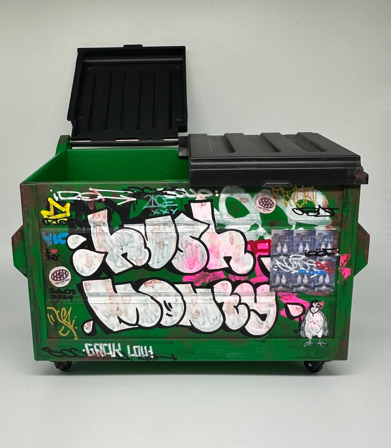 May include: A green metal dumpster with a black lid. The dumpster is covered in graffiti, including the words "Crush It" and "Go Big Lou!"