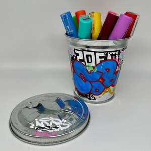 May include: A silver metal trash can with a graffiti design featuring blue, yellow, red, and green paint. The can has a lid and is filled with colorful markers.