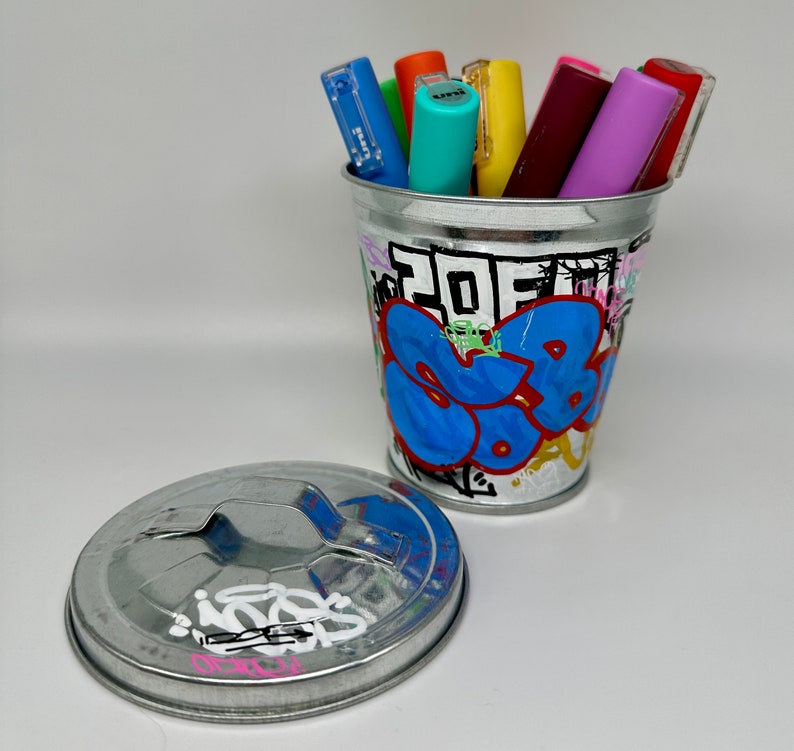 May include: A silver metal trash can with a graffiti design featuring blue, yellow, red, and green paint. The can has a lid and is filled with colorful markers.