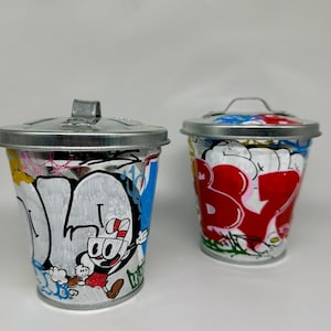 May include: Two small metal trash cans with lids. One is painted with a cartoon character and the other is painted with colorful graffiti.