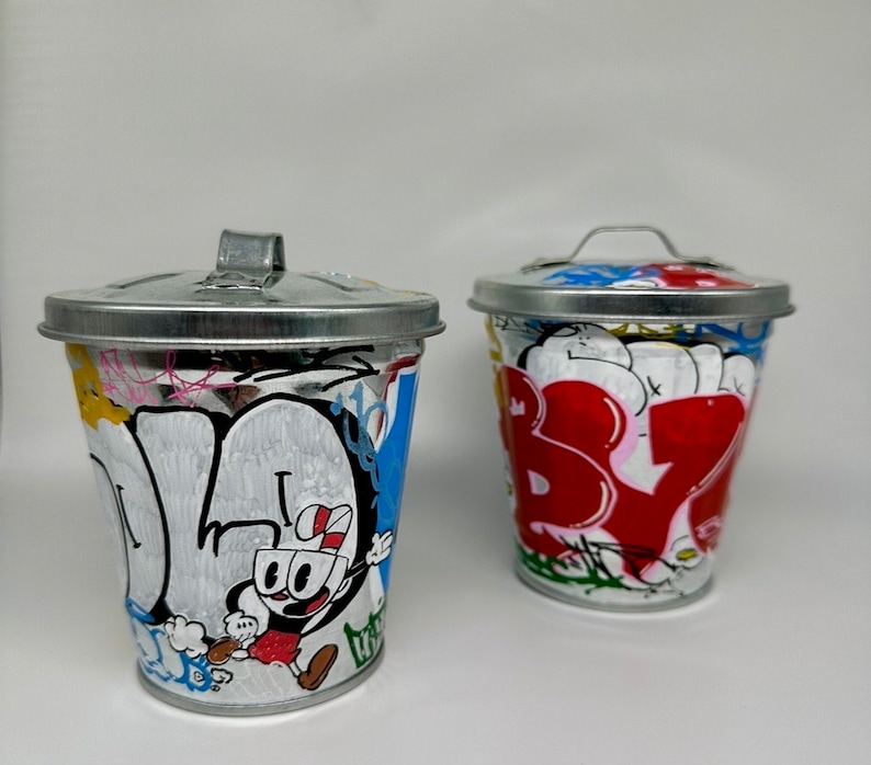 May include: Two small metal trash cans with lids. One is painted with a cartoon character and the other is painted with colorful graffiti.
