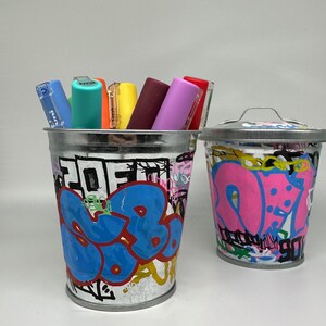 May include: Two metal containers with graffiti-style designs. One container is pink and white and the other is blue, white, and black. Both containers are filled with colorful markers.