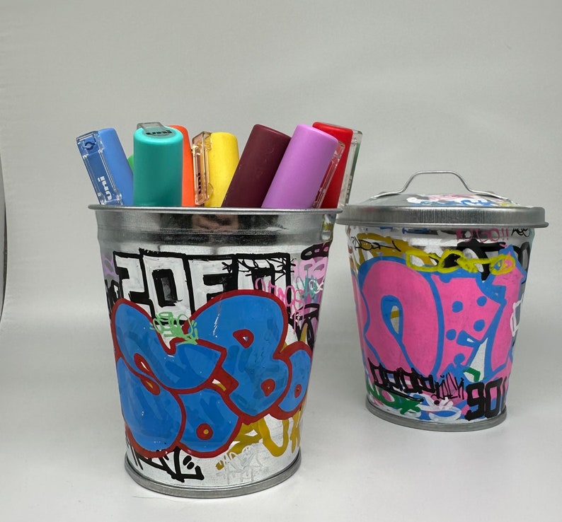 May include: Two metal containers with graffiti-style designs. One container is pink and white and the other is blue, white, and black. Both containers are filled with colorful markers.