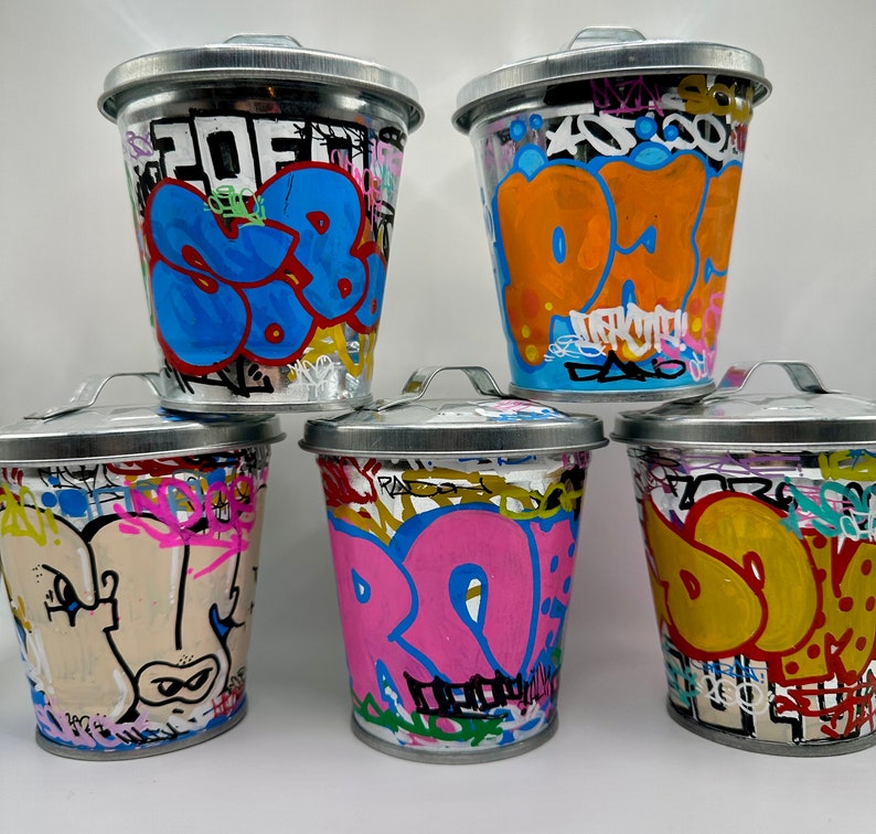 May include: Five metal trash cans with colorful graffiti designs. The cans are all different colors and have different designs. The designs include words, letters, and abstract shapes.