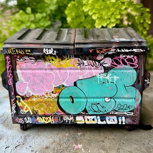 Op de afbeelding: Een miniatuur vuilnisbak met een zwart deksel en zijkanten, bedekt met kleurrijke graffiti. De graffiti bevat roze, gele en turquoise ontwerpen, samen met verschillende tags en het nummer "2024". De vuilnisbak heeft kleine wielen.