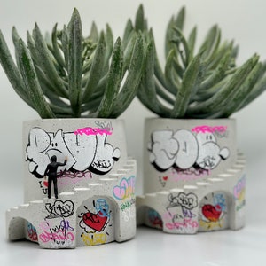 May include: Two gray concrete planters shaped like castles with graffiti designs. Each planter has a succulent plant with green leaves. One planter has a small black figure painting graffiti on the side.