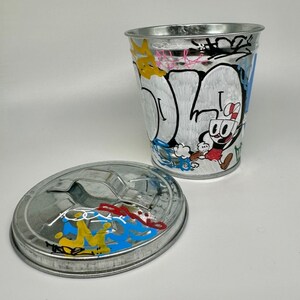 May include: A silver metal trash can with a lid. The can is decorated with colorful graffiti, including a cartoon character. The lid is also decorated with graffiti.