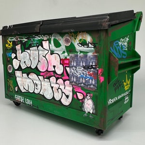 May include: A green metal dumpster with graffiti art. The graffiti includes the words "Crush" and "Nobody" in pink and white letters. There is also a sticker with a bird and the words "Phil Rules" and "Happy Birthday".