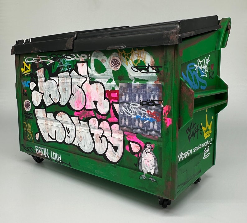 May include: A green metal dumpster with graffiti art. The graffiti includes the words "Crush" and "Nobody" in pink and white letters. There is also a sticker with a bird and the words "Phil Rules" and "Happy Birthday".