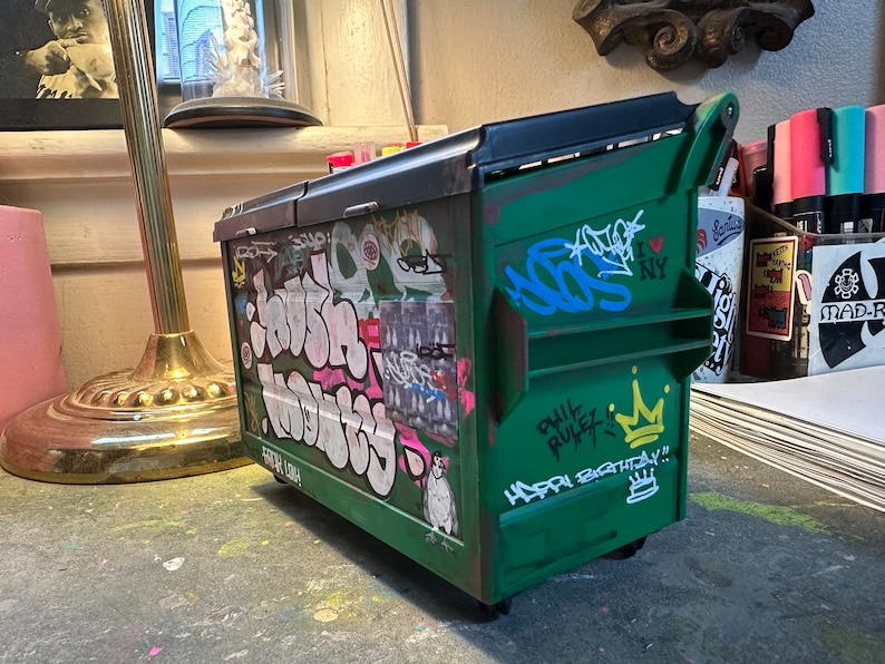 May include: A green metal trash can with a black lid and graffiti on the sides. The graffiti includes the words "Phil Rulez", "Happy Birthday", and "I Love NY".