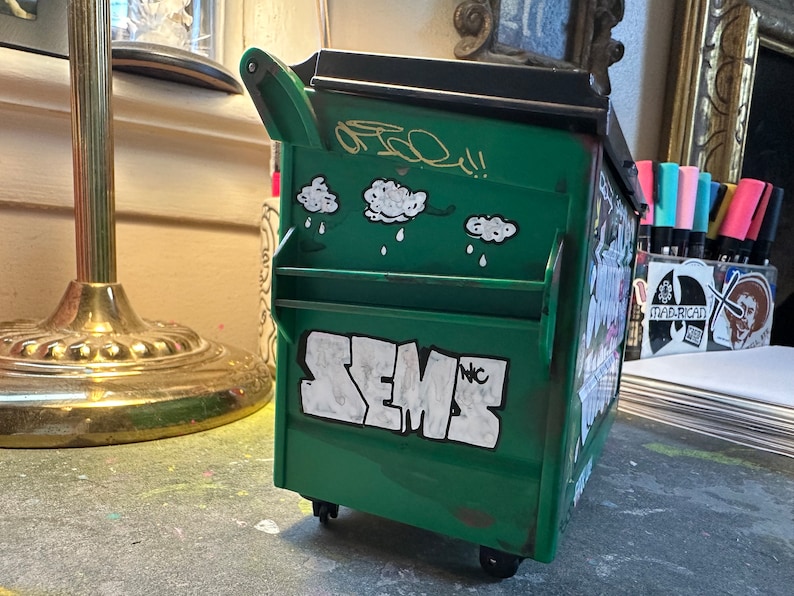 May include: A green toy dumpster with a black lid and wheels. The dumpster has white graffiti that says "SEMS" and "NYC". There are also three white cloud stickers with raindrops on the side of the dumpster.