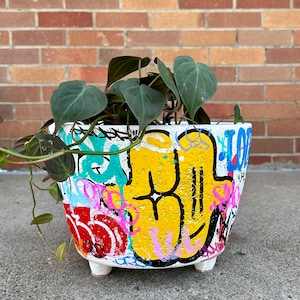 Op de afbeelding: Witte plantenpot versierd met graffiti-kunst in diverse kleuren, waaronder geel, blauw, roze en rood. De pot bevat een groene plant met bladeren. De pot heeft drie kleine, witte voetjes.