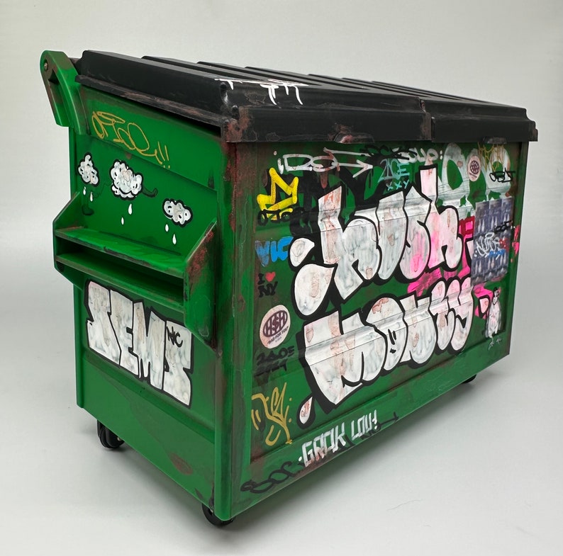 May include: A green metal dumpster with graffiti art. The graffiti includes the words "Crush" and "Semc" in large letters, as well as other smaller words and designs. The dumpster has a black lid and a handle on the side.
