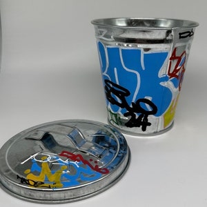 May include: A small, silver metal trash can with a lid. The can is decorated with colorful graffiti, including blue, yellow, red, and black paint. The lid has a handle.