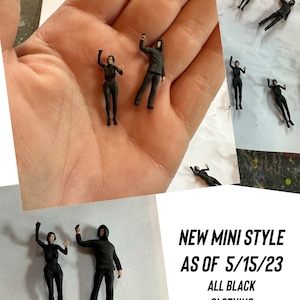 May include: Two small black figures, a male and a female, wearing all black clothing, are posed with their arms raised. The figures are standing on a hand. The text "NEW MINI STYLE AS OF 5/15/23 ALL BLACK CLOTHING" is visible in the image.