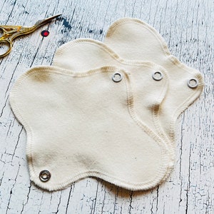 Könnte beinhalten: Drei weiße wiederverwendbare Stoff-Menstruationseinlagen mit Druckknöpfen. Die Einlagen sind übereinander gefaltet und liegen auf einer weißen Holzoberfläche.