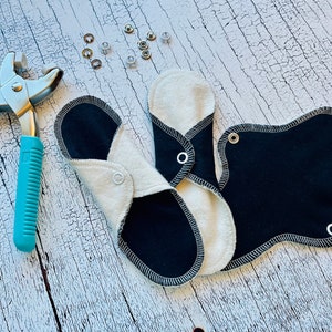 Peut inclure: Trois coussinets menstruels r&eacute;utilisables en tissu noir et blanc avec une fermeture &agrave; pression. Un outil argent&eacute; avec une poign&eacute;e bleue est utilis&eacute; pour fixer les pressions.