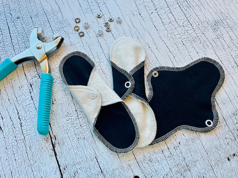 Peut inclure: Trois coussinets menstruels r&eacute;utilisables en tissu noir et blanc avec une fermeture &agrave; pression. Un outil argent&eacute; avec une poign&eacute;e bleue est utilis&eacute; pour fixer les pressions.