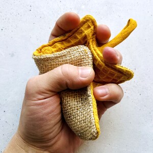 May include: A hand holding a yellow waffle weave dishcloth with a burlap backing. The dishcloth is folded in half and the hand is squeezing it.