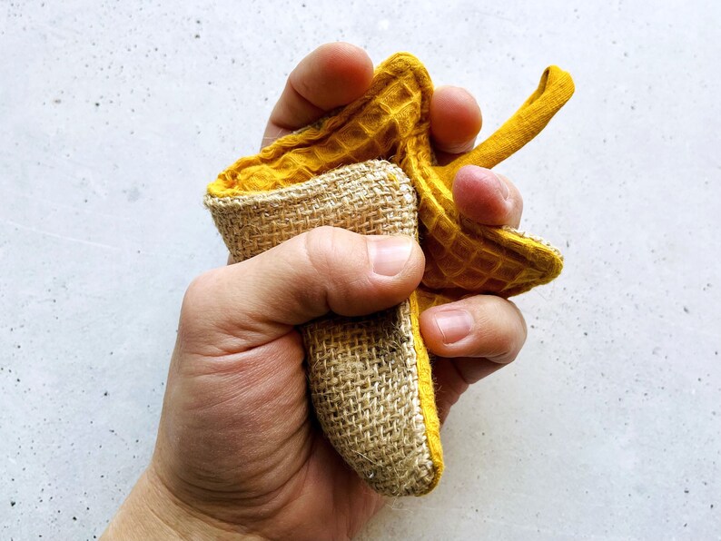 May include: A hand holding a yellow waffle weave dishcloth with a burlap backing. The dishcloth is folded in half and the hand is squeezing it.