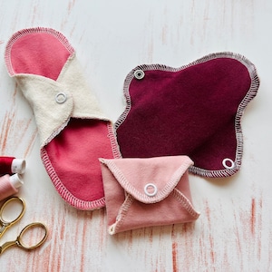 Peut inclure: Trois serviettes menstruelles en tissu r&eacute;utilisables de diff&eacute;rentes couleurs et tailles. Les serviettes sont pli&eacute;es et ont des boutons pression sur les c&ocirc;t&eacute;s. Les serviettes sont sur une surface en bois blanc.