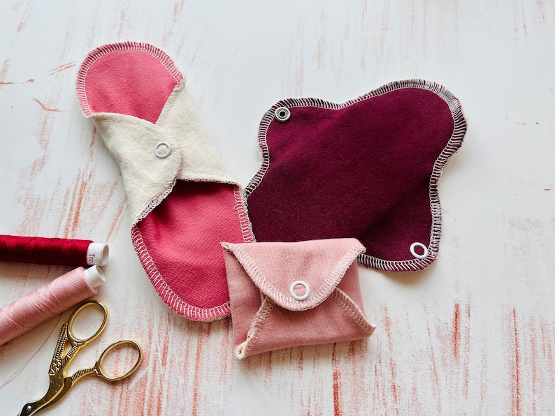 Peut inclure: Trois serviettes menstruelles en tissu r&eacute;utilisables de diff&eacute;rentes couleurs et tailles. Les serviettes sont pli&eacute;es et ont des boutons pression sur les c&ocirc;t&eacute;s. Les serviettes sont sur une surface en bois blanc.