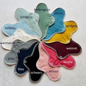 Peut inclure: Un nuancier de 12 coussinets lavables en tissu de diff&eacute;rentes couleurs, dont vert clair, p&eacute;trole, jaune moutarde, rouge vin, corail et noir. Les coussinets sont dispos&eacute;s en cercle avec les noms des couleurs &eacute;crits en allemand.