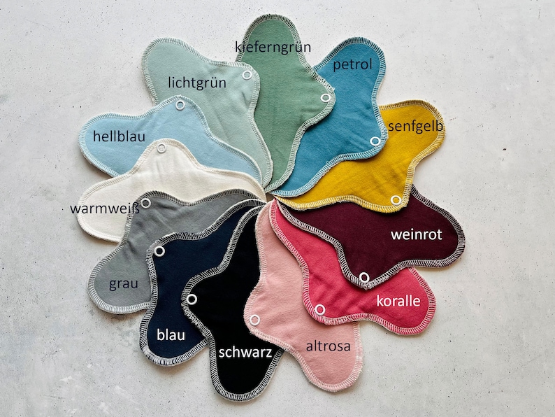 Peut inclure: Un nuancier de 12 coussinets lavables en tissu de diff&eacute;rentes couleurs, dont vert clair, p&eacute;trole, jaune moutarde, rouge vin, corail et noir. Les coussinets sont dispos&eacute;s en cercle avec les noms des couleurs &eacute;crits en allemand.
