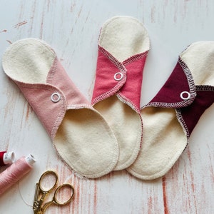 Peut inclure: Trois serviettes menstruelles en tissu r&eacute;utilisables de couleur rose, rouge et bordeaux avec des boutons-pression blancs. Les serviettes sont pli&eacute;es et dispos&eacute;es sur une surface en bois blanc. On voit &eacute;galement deux bobines de fil et une paire de ciseaux dor&eacute;s sur l'image.