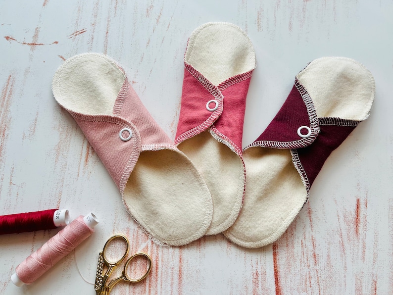 Peut inclure: Trois serviettes menstruelles en tissu r&eacute;utilisables de couleur rose, rouge et bordeaux avec des boutons-pression blancs. Les serviettes sont pli&eacute;es et dispos&eacute;es sur une surface en bois blanc. On voit &eacute;galement deux bobines de fil et une paire de ciseaux dor&eacute;s sur l'image.