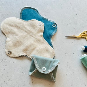 Peut inclure: Trois coussinets menstruels r&eacute;utilisables en tissu de diff&eacute;rentes nuances de bleu et beige. Les coussinets sont dot&eacute;s de boutons-pression et sont dispos&eacute;s sur une surface blanche avec du fil et des ciseaux.