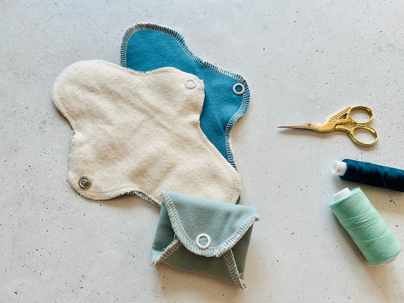 Peut inclure: Trois coussinets menstruels r&eacute;utilisables en tissu de diff&eacute;rentes nuances de bleu et beige. Les coussinets sont dot&eacute;s de boutons-pression et sont dispos&eacute;s sur une surface blanche avec du fil et des ciseaux.