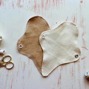 Könnte beinhalten: Zwei wiederverwendbare Stoff-Menstruationseinlagen, eine braune und eine weiße, mit weißen Druckknöpfen. Die Einlagen liegen auf einer weißen Holzoberfläche.