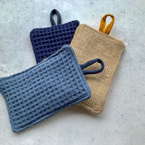 May include: Three kitchen scrubbers, one is a dark blue waffle weave with a loop handle, another is a light blue waffle weave with a loop handle, and the last is a natural burlap scrubber with a yellow loop handle.