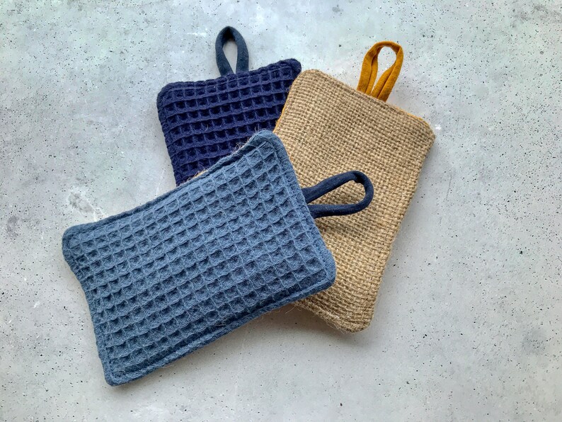 May include: Three kitchen scrubbers, one is a dark blue waffle weave with a loop handle, another is a light blue waffle weave with a loop handle, and the last is a natural burlap scrubber with a yellow loop handle.