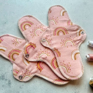 Könnte beinhalten: Drei wiederverwendbare Stoff-Menstruationseinlagen in Pink mit einem Regenbogen- und Goldpunktmuster. Die Einlagen haben Druckknöpfe an den Seiten.