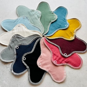 Peut inclure: Un ensemble de 12 serviettes menstruelles en tissu r&eacute;utilisables de diff&eacute;rentes couleurs, notamment vert, bleu, jaune, rouge, rose, gris et noir. Les serviettes sont dispos&eacute;es en cercle, les boutons pression vers l'ext&eacute;rieur.