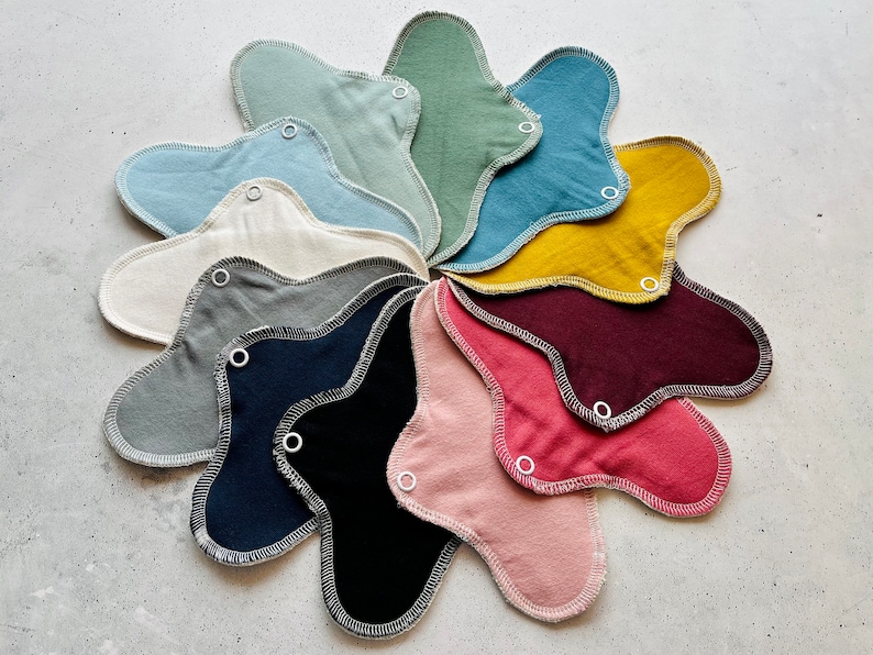 Peut inclure: Un ensemble de 12 serviettes menstruelles en tissu r&eacute;utilisables de diff&eacute;rentes couleurs, notamment vert, bleu, jaune, rouge, rose, gris et noir. Les serviettes sont dispos&eacute;es en cercle, les boutons pression vers l'ext&eacute;rieur.