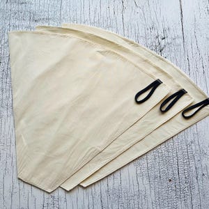Könnte beinhalten: Drei kegelförmige Kaffeefilter aus cremefarbenem Stoff. Jeder Filter verfügt über eine genähte Kante und eine kleine schwarze Stoffschlaufe an der Seite zum Aufhängen. Die Filter sind fächerförmig übereinander angeordnet.