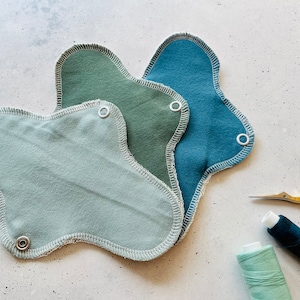 Peut inclure: Trois serviettes menstruelles en tissu r&eacute;utilisables dans diff&eacute;rentes nuances de vert et de bleu. Les serviettes sont pli&eacute;es et empil&eacute;es les unes sur les autres. Il y a une bobine de fil vert et une bobine de fil bleu, ainsi qu'une paire de ciseaux dor&eacute;s sur une surface blanche.