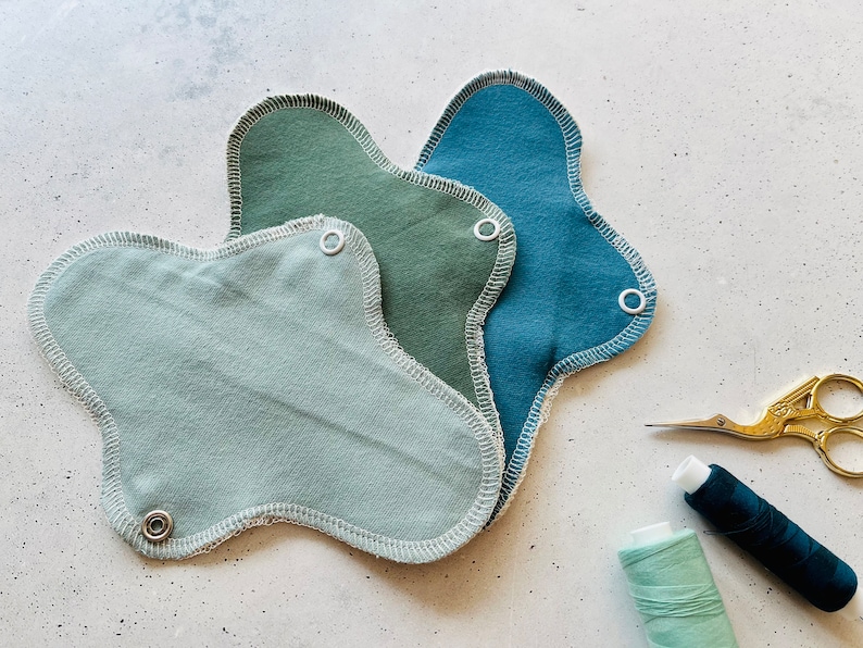Peut inclure: Trois serviettes menstruelles en tissu r&eacute;utilisables dans diff&eacute;rentes nuances de vert et de bleu. Les serviettes sont pli&eacute;es et empil&eacute;es les unes sur les autres. Il y a une bobine de fil vert et une bobine de fil bleu, ainsi qu'une paire de ciseaux dor&eacute;s sur une surface blanche.