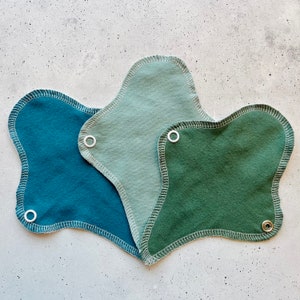 Könnte beinhalten: Drei wiederverwendbare Stoff-Menstruationseinlagen in verschiedenen Grüntönen. Die Einlagen sind aus weichem Stoff und haben weiße Nähte an den Rändern. Jede Einlage hat einen weißen Druckknopf an der Seite.