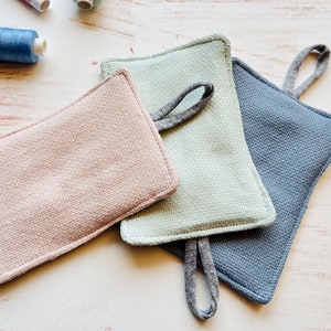 Puede incluir: Tres agarraderas rectangulares texturizadas, rosa, verde y azul, cada una con un lazo para colgar. Están dispuestas sobre una superficie clara, con carretes de hilo en el fondo.