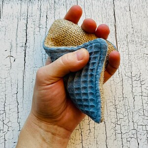 May include: A hand holds a blue and brown textured cleaning sponge. The sponge is made of natural materials and is designed for cleaning dishes and other surfaces.