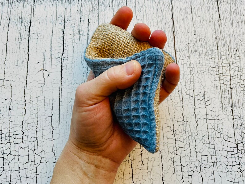 May include: A hand holds a blue and brown textured cleaning sponge. The sponge is made of natural materials and is designed for cleaning dishes and other surfaces.