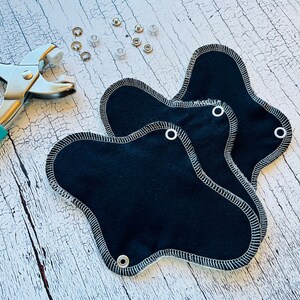 Peut inclure: Trois serviettes hygi&eacute;niques menstruelles en tissu noir avec des coutures blanches et des boutons-pression m&eacute;talliques. Un outil m&eacute;tallique pour fixer les boutons-pression est &eacute;galement illustr&eacute;.