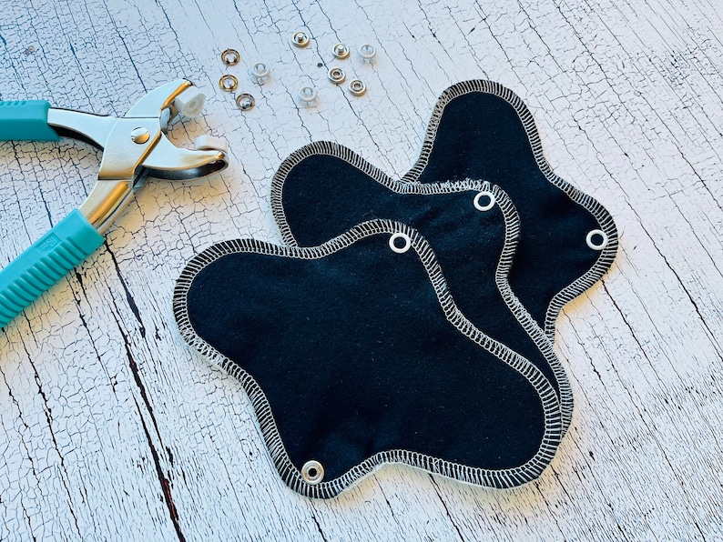 Peut inclure: Trois serviettes hygi&eacute;niques menstruelles en tissu noir avec des coutures blanches et des boutons-pression m&eacute;talliques. Un outil m&eacute;tallique pour fixer les boutons-pression est &eacute;galement illustr&eacute;.